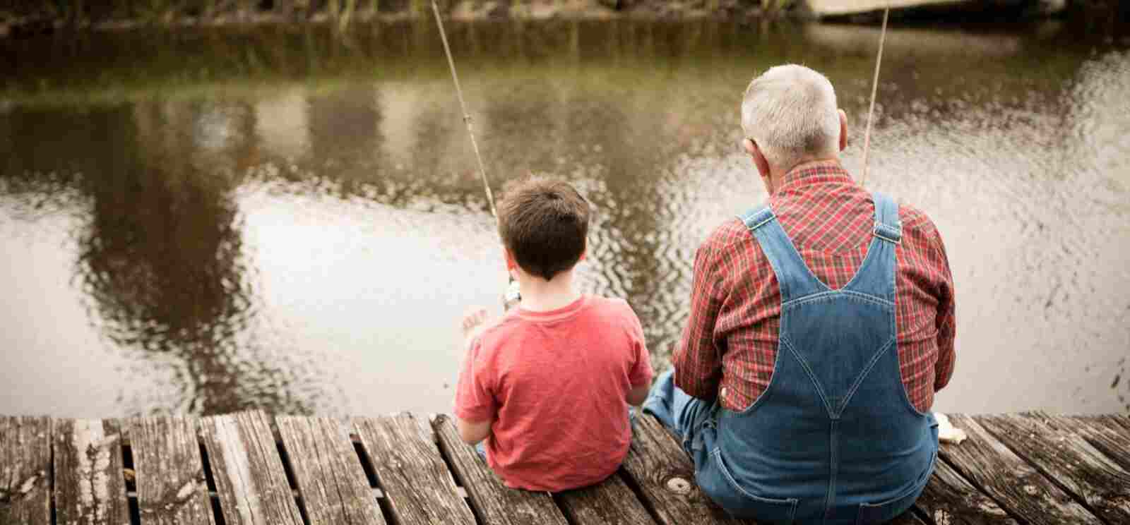 Grandpa and grandson sitting on the edge of a pier overlooking a pond holding fishing poles