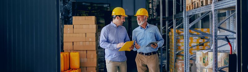 Two men in hard hats walk through warehouse.