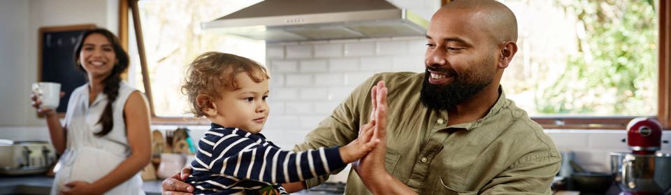 Pregnant woman watches from behind as her husband gives high five to young child.