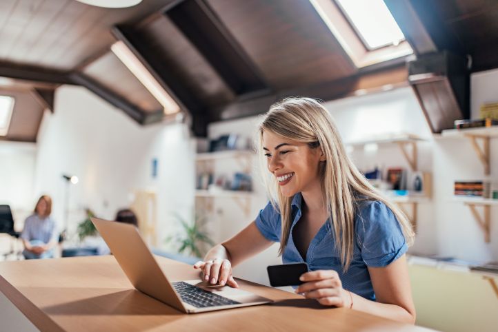 Woman sitting at a desk smiling, working on a laptop and holding a credit card.