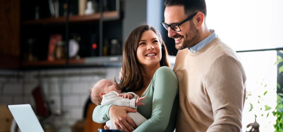 A young couple holding a newborn baby and smiling at each other at home. 