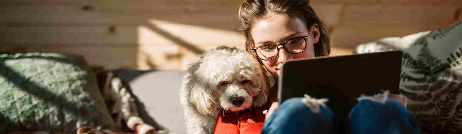 Woman holding laptop sits next to dog on couch.