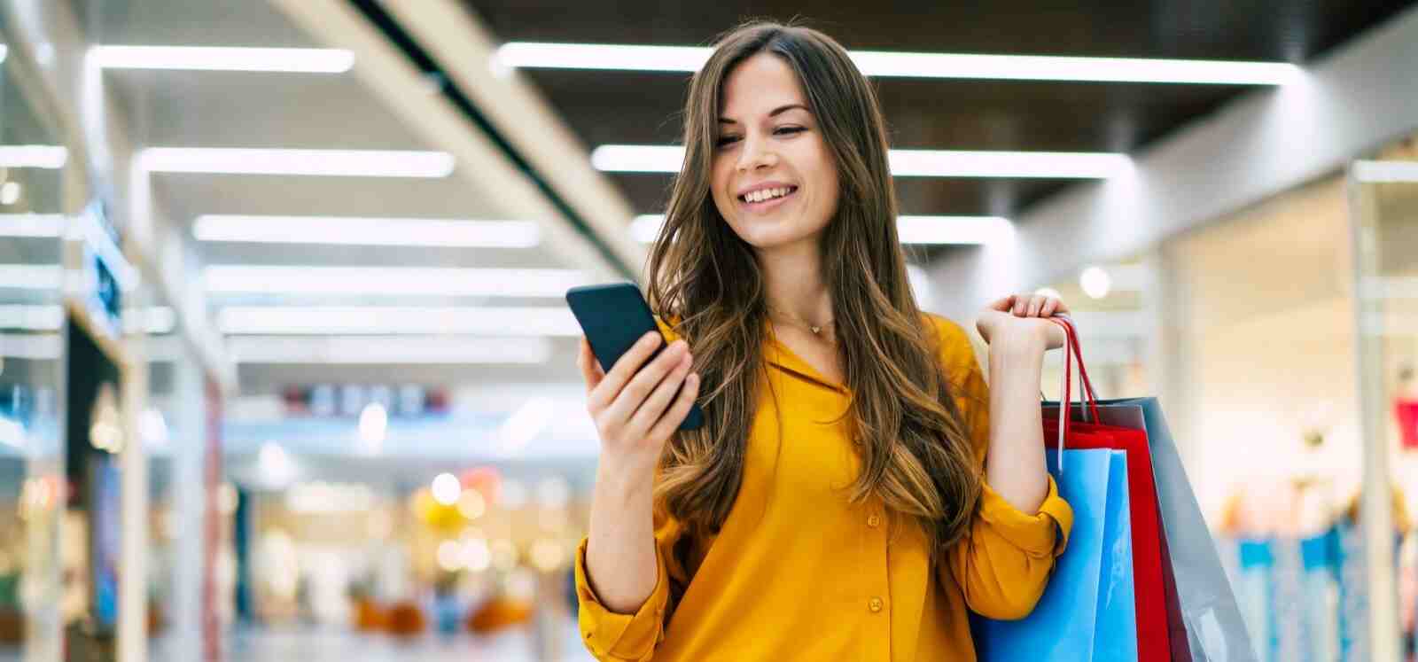 woman shopping at a mall holding bags and looking at her phone smiling